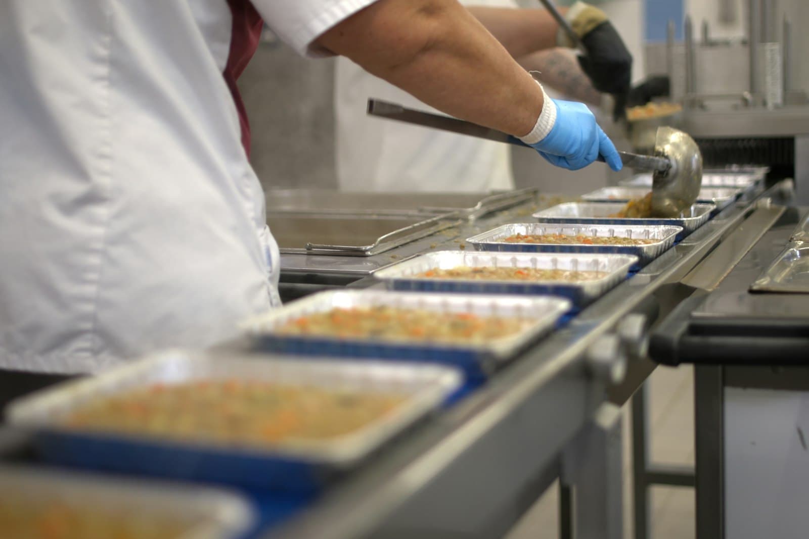 A worker in gloves portioning food into trays moving along a conveyor line